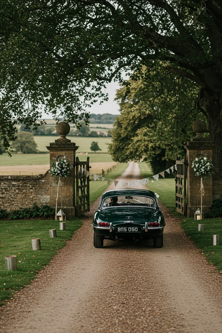 A vintage green car with ‘Just Married’ bunting drives out between stone gateposts, towards open fields.