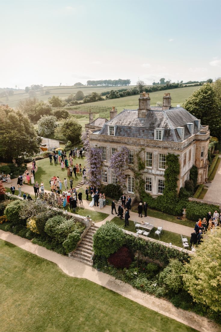 Ashden Manor from above: a pale stone Georgian house with wisteria on its walls, wedding guests gathered on the lawn and fields rolling away behind.