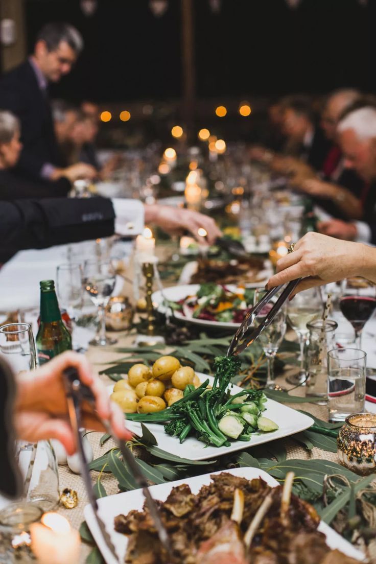 Guests help themselves from platters of new potatoes, tenderstem broccoli and roast meat along a long candlelit table.